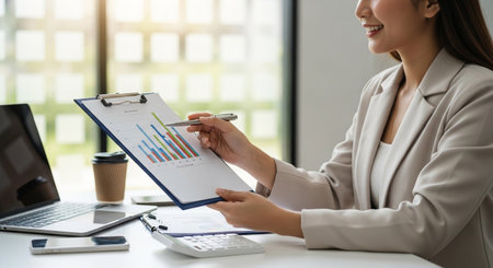 A focused businesswoman analyzes financial data on a clipboard, using a pen to highlight key points of a bar graph in a modern office setting.の素材
