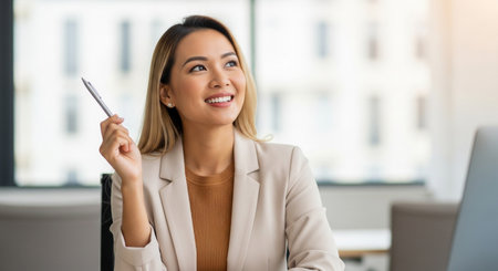 A professional woman in a business suit smiles while holding a pen, looking upwards with a thoughtful expression, suggesting contemplation and future planning.の素材