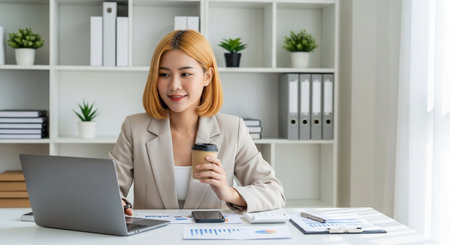 A focused Asian professional enjoys a coffee break while diligently working on her laptop, embodying modern workplace efficiency and ambition.の素材