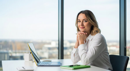 A confident businesswoman smiles warmly while seated at her desk, a laptop open before her, in a contemporary office space offering a panoramic view of the cityscape through large windows.の素材