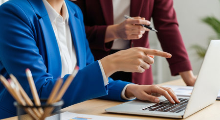 In a bright office, two businesswomen work together. One points at the laptop screen, guiding the other who is typing. Pencils rest nearby, ready for note-taking and brainstorming.の素材