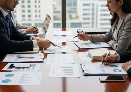 A group of business professionals are gathered around a table, reviewing financial reports and charts, pointing at data, and discussing strategies for business growth and success.の素材