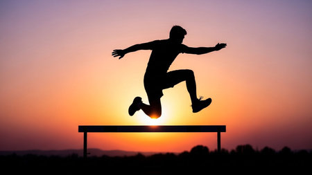 A silhouette of a male athlete mid-jump over a hurdle against a vibrant sunset sky.の素材