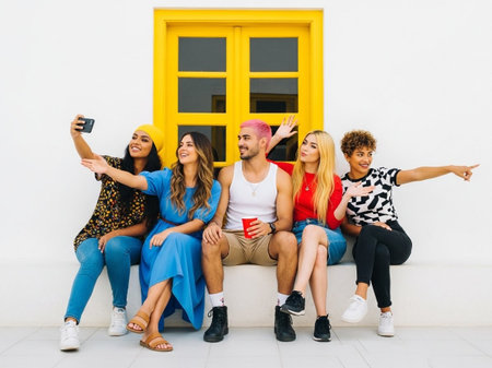 Six young people sit on a bench in front of a bright yellow window, wearing casual clothing and smiling while taking a selfieの素材