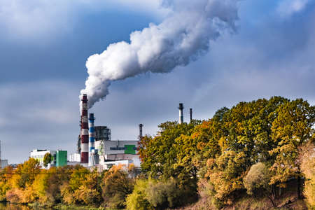 pipes of woodworking enterprise plant sawmill near river with autumn red yellow trees. Air pollution concept. Industrial landscape environmental pollution waste of thermal power plantの写真素材