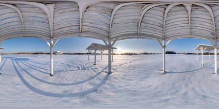 Winter full spherical seamless panorama 360 degrees angle view snow covered deserted beach with gazebos near lake  in snowy park with blue sky at evening in equirectangular projection. VR AR contentの写真素材