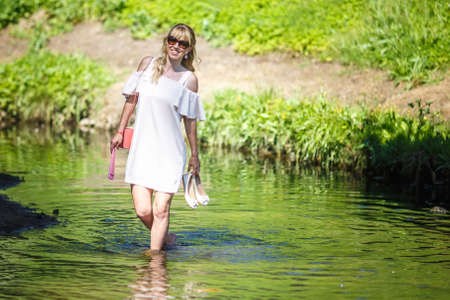 beautiful girl walks on river water with shoes in hands in sunglasses