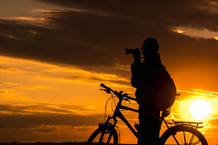Silhouette of male photographer cyclist stands on the road and looks into the distance and admires the beautiful landscape at crimson sunset.の写真素材