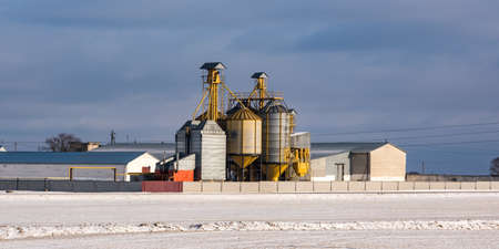 agro silos granary elevator in winter day in snowy field. Silos on agro-processing manufacturing plant for processing drying cleaning and storage of agricultural products, flour, cereals and grain.の写真素材