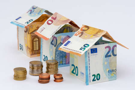 Houses made of 10, 20 and 50 euro bank notes, stacks of euro coins, white background. Concepts of prices and costs linked to housing and real estateの写真素材