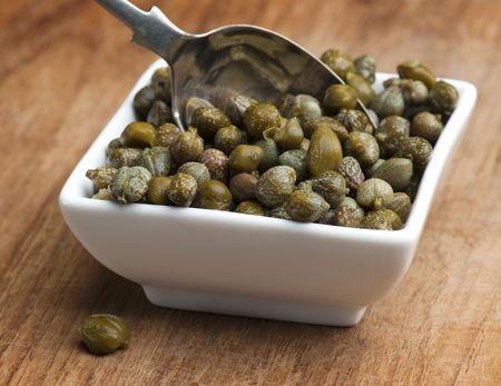 A White Dish Full Of Capers, With A Spoon Being Put In To Them, Up On A Wooden Surface