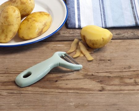 A Potato Peeler With Potatoes In An Enamel Dish and A Cloth In The Backgroundの写真素材