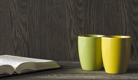 Two ceramic mugs yellow and green stand in the corner of a wooden table in front of an open bookの写真素材