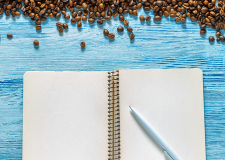 Single simple empty white notebook with a blank for drawing or writing, white plastic pen and scattered roasted coffee beans are on a Desktop from blue wooden plank. Top view. Mock up. Flat layの写真素材