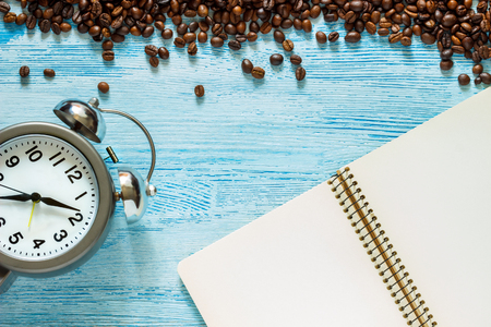 Single simple empty white notebook with a blank for drawing or writing, scattered roasted coffee beans and alarm clock with silver metal bells are on a Desktop from blue wooden plank. Top view. Mock up. Flat layの写真素材