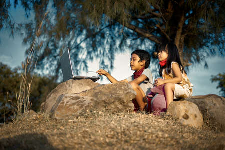 A boy and his little cute sister sitting on a rock and study using laptops under the tree.  New normal for Education conceptの写真素材