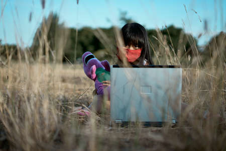 Cute little girl with protected mask and using laptop sitting on grass, New normal of Education...