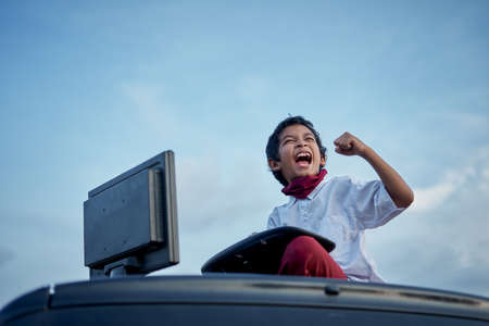 Excited Schoolboy fist pumps while working on a homework using computer on top of car roof, New Normal Education conceptの写真素材