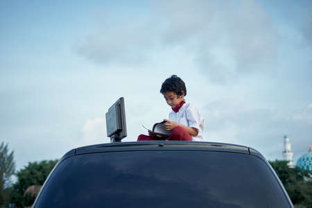 Schoolboy doing homework using computer on top of car roof, New Normal Education concept