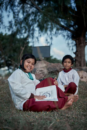 Two happy elementary students with a book and computer studying at natural park, New Normal Education conceptの写真素材
