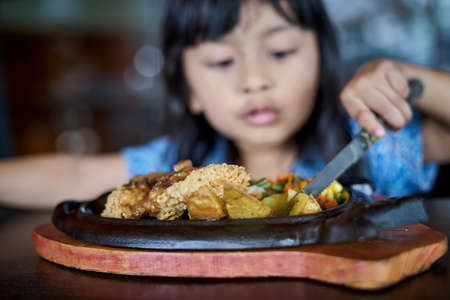 Little girl holding knife and fork cutting grilled Chicken steak on stoned plate in restaurantの写真素材