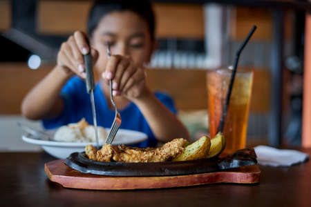 Boy holding knife and fork cutting grilled Chicken steak on stoned plate in restaurantの写真素材