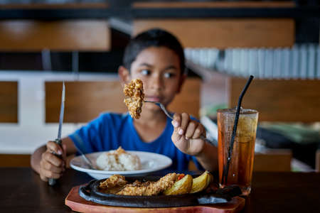 Boy holding knife and fork cutting grilled Chicken steak on stoned plate in restaurantの写真素材