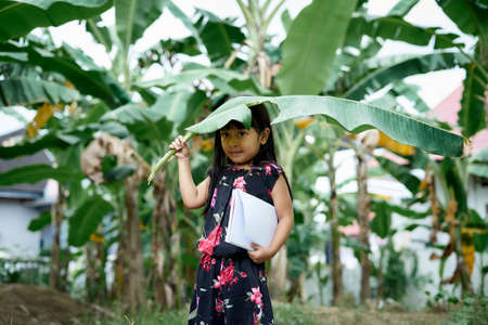 Little girl with banana leaf umbrella and holding a note book playing at parkの写真素材