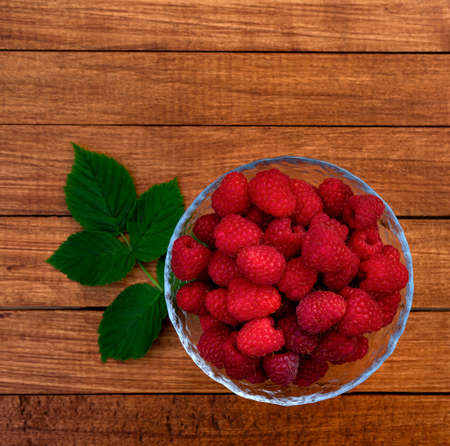 Ripe large juicy raspberries in a transparent plate on a wooden table with green leaves.