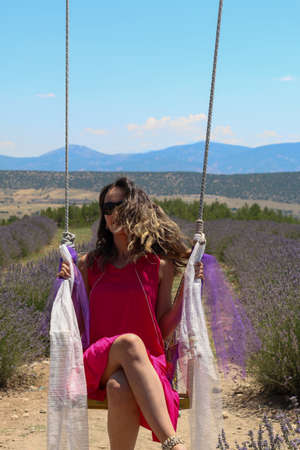 Turkish woman in brunette pink dress with brunette sunglasses sitting on swing in lavender field under sky, copy spaceの写真素材
