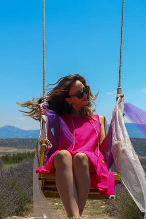 Turkish woman in brunette pink dress with brunette sunglasses sitting on swing in lavender field...