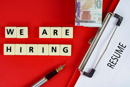 Business concept - layout of fake money, pen, resume on clipboard and tiles with text We are hiringの写真素材