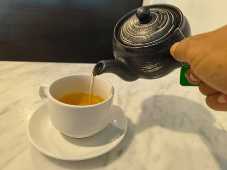 Pouring hot green tea from a dark, rustic Japanese clay teapot into a smooth, white porcelain cup. The warm steam rises as the tea flows in a delicate stream, creating a calming, traditional scene.の写真素材