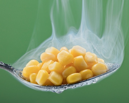 Macro photo of preserved corn vegetable in metal spoon on a light green background with steam.