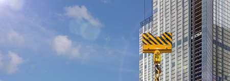 Construction scene - a glass skyscraper under construction against a blue sky and a yellow crane hook in the foreground. Blank for an advertising banner about real estate or construction. Copy spaceの写真素材