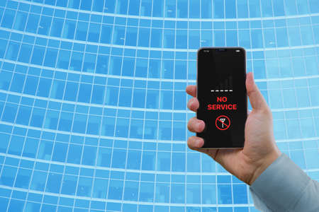 A mans hand holds a smartphone with the notification No service against the background of a business building with a blue glass facade. Notification No service on the smartphone screenの写真素材