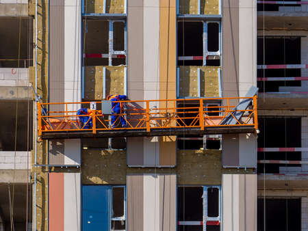 The process of insulating external walls in a multi-storey building under construction. Workers in an orange construction cradle attach mineral wool sheetsの写真素材