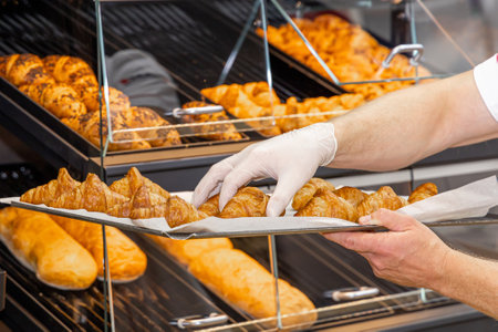 A baker places freshly baked croissants on shelves in a bakery. Production and sale of bread, croissants and loaves. Fresh pastries, bakery products. Selected focus.の写真素材