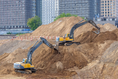 Two yellow crawler excavators on a construction site - one on the lower level, the other on top of a...