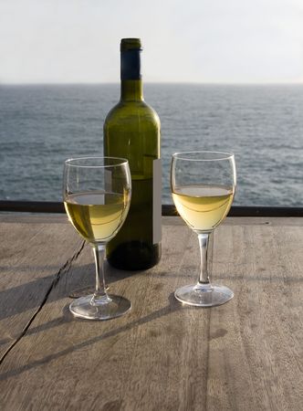 Two glasses with white wine and a bottle on a wooden table with view on ocean in early setting sunの写真素材