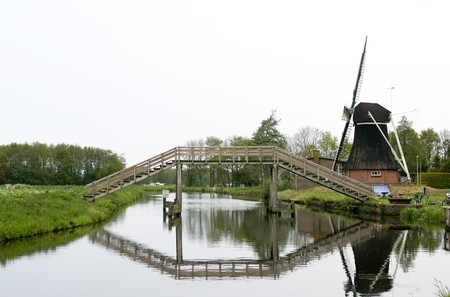 Typical Dutch wooden high bridge above river with windmill and reflection of bridge in waterの写真素材