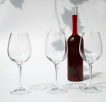 three empty wine glasses and a bottle filled with red wine against a background with the shadows of vine leavesの写真素材