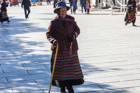 Lhasa, Tibet, China - November 15, 2019: Old Tibetan woman  on  crutches on the Jokhang Temple Square.  Poor looking elderly woman in traditional Tibetan clothing and blue hatのeditorial素材