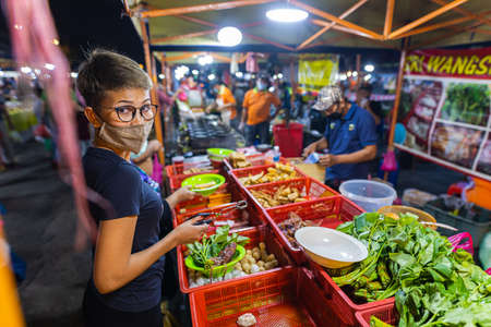 Street food night market Putrajaya, near Kuala Lumpur. Young asian girl buys vegetables in a night market. Malaysian women with face mask in a street marketのeditorial素材