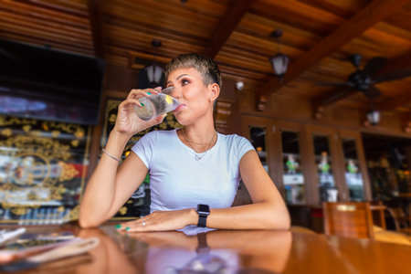 A young asian girl sitting alone at a wooden table in a restaurant and drinking a glass of water. Portrait of a pretty smiling Malaysian woman, sitting at a table in a cafÃ© in Kuala Lumpur, Malaysia.の写真素材