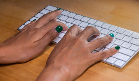 Woman hands typing on a computer keyboard. Female hands writing on a aluminum computer keyboard. Fingernails are painted in green. the desk is made of wood. Home office during COVID lockdownのeditorial素材