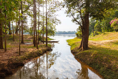 Traditional long tail wooden boat in a small lake near Siem Reap, Cambodia. The sky and the trees around is reflected in the water. Idyllic place just near the most visited place of Cambodiaの写真素材