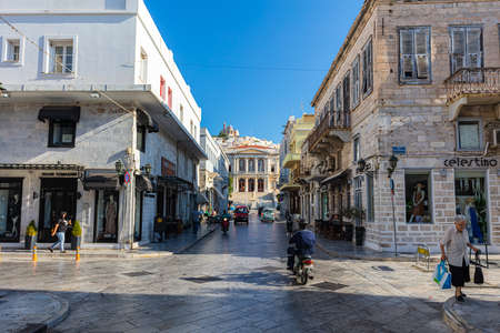Syros, Greece - July 1, 2021: View into the narrow alley near the town hall and Miaouli square at the island of Syros. Located in the Aegean Sea in the cyclades archipelago. People walking on sidewalkのeditorial素材