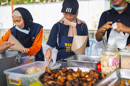 Kuala Lumpur, Malaysia - June 21, 2022: Young girls selling bbq chicken on a street market. Marinated grilled chicken are put in a paper bag by two teenage Muslim women. Chicken seller at night marketのeditorial素材