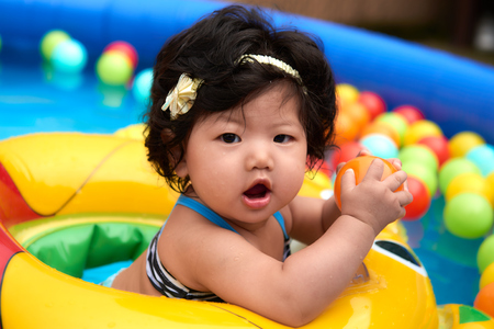 Asian baby girl in swimsuit and float swimming in pool with ballsの写真素材